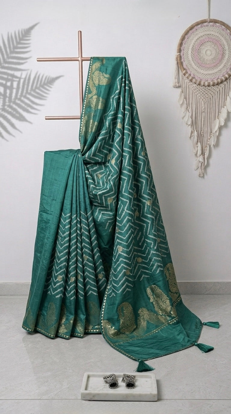 A teal green saree with gold zari patterns draped over a wooden stand in a styled room setting, with a matching folded section beside it, decorative wall hanging, and a small tray on the floor.