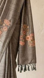 A draped gray sari with peach and pink floral embroidery and green tassel accents, hanging from a rustic wooden branch against a warm beige wall. A small wooden stool with a black clutch is visible in the lower left corner.